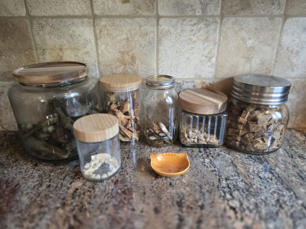 Glass jars filled with natural dog and cat treats stored on a kitchen counter, replacing plastic packaging for cleaner pet food storage.