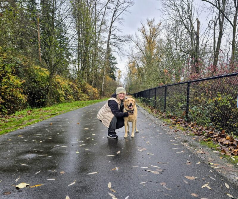 Woman crouching beside a yellow Labrador on a wet fall trail.
